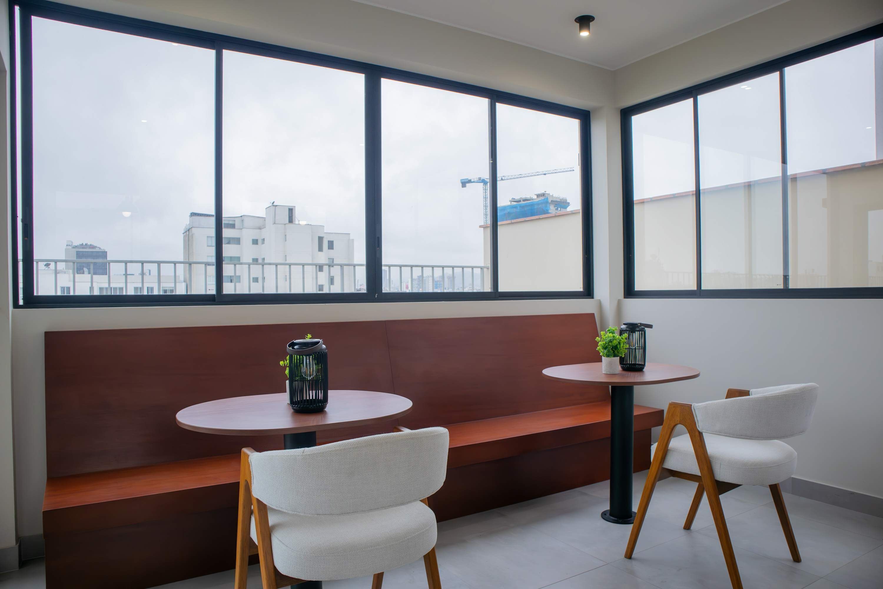 A modern interior with two round tables, white chairs, and a wooden bench. Large windows offer a view of the cityscape.