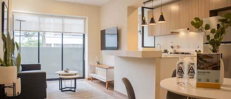 A modern living room with a sofa, coffee table, and a dining area with chairs and a table. There is a large window providing natural light and a view outside.