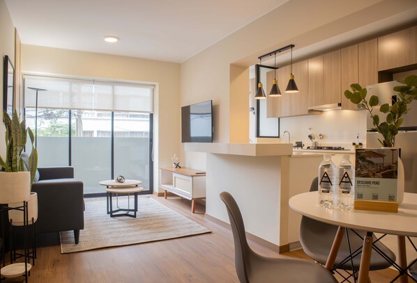 A modern living room with a sofa, coffee table, and a dining area with chairs and a table. There is a large window providing natural light and a view outside.