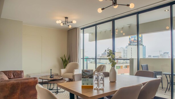 A modern hotel room with a large window, a wooden dining table, comfortable seating, and a view of the cityscape.