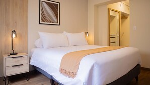 A neatly made bed with white linens and a beige throw blanket, a bedside table with a lamp, and a framed picture on the wall.