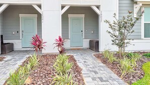 A modern building with two light blue doors, a covered patio area with a brick pathway, and various plants. - 16037 Dream Vacation Family Home 5BD (Clermont)