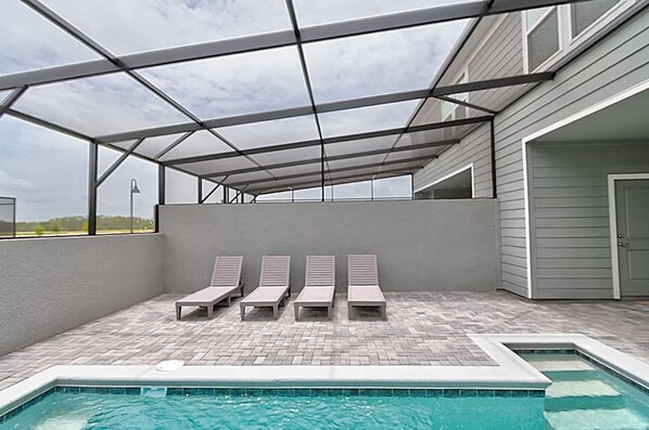 Una piscina exterior con un porche cubierto, cuatro sillas de descanso y una vista despejada del cielo.