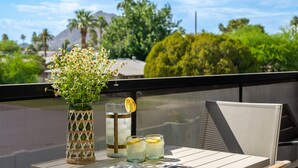 A table with a vase of flowers, two glasses of iced drinks with lemon slices, and a chair on an outdoor balcony with a view of greenery and mountains. - NEW Desert Standard Modern 3-level Oasis (Scottsdale)
