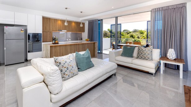 A modern living room with a white sofa and a smaller sofa, both with patterned cushions. A kitchen with wooden cabinets and stainless steel appliances is visible in the background. - 3BR Lux Beachfront Escape - Pool Gym King bed (Sunshine Coast)