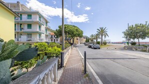 A coastal street with multi-story buildings, a sidewalk, and parked cars. - Next to the Beach by Wonderful Italy (Ospedaletti)