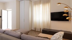 Una habitación de hotel moderna con sofá, televisor montado en la pared, armario y un ventanal con cortinas.