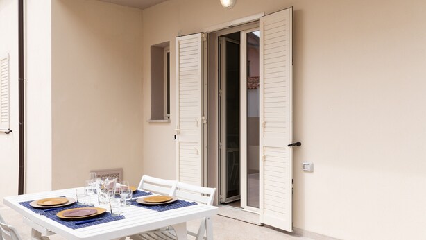 Un comedor con una mesa lista para cuatro personas, sillas blancas y un ventanal corredero que da a un espacio exterior.