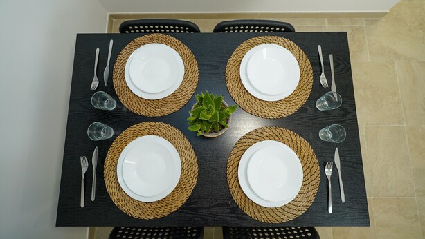 Une table de salle à manger rectangulaire, sur laquelle sont disposées quatre assiettes blanches, chacune entourée d’un set de table en fibres tressées, avec couverts et verres.