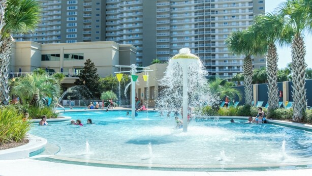 A swimming pool with a central fountain, surrounded by palm trees and a multi-story building in the background.