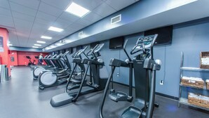 A gym with multiple treadmills and ellipticals, a red wall, and a shelf with baskets on the right.
