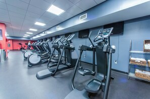 Ein Fitnessraum mit mehreren Laufbändern und Crosstrainern, einer roten Wand und einem Regal mit Körben auf der rechten Seite.