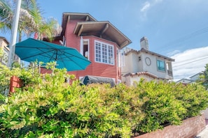 A red house with a balcony, a green umbrella, and lush greenery in the foreground.