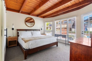 A bedroom with a wooden ceiling, a bed, a nightstand, a desk, and a large window with blinds.