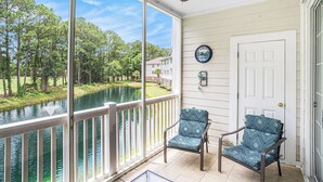 A covered porch with a view of a lake and trees, two patterned chairs, a ceiling fan, and a door with a clock.