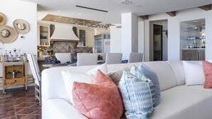 Ein modernes Wohnzimmer mit einem weißen Sofa, bunten Kissen, einem Holztisch und einer Küche im Hintergrund.