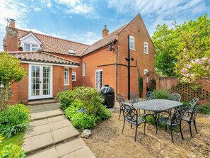 A brick house with a tiled roof, a covered patio with metal chairs and a table, and a garden with various plants.