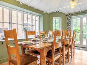 A dining area with a wooden table set for six, surrounded by chairs, with windows and a door in the background.