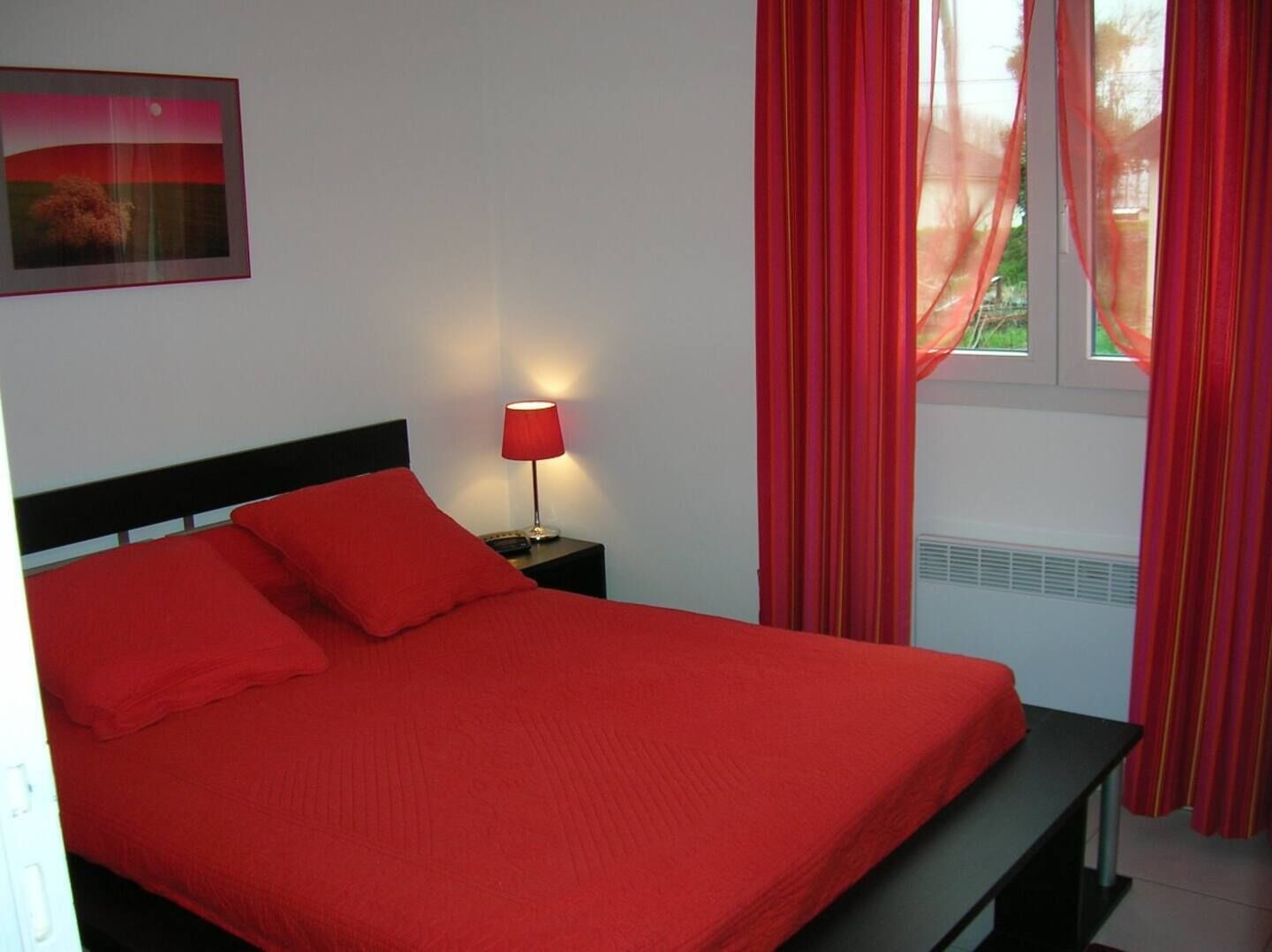 Una cama con ropa de cama roja, una lámpara de noche y una ventana con cortinas rojas.