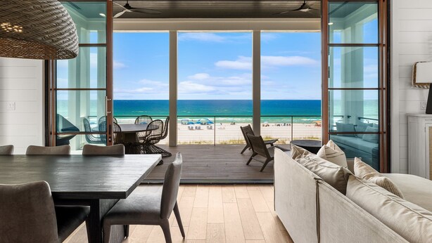 A modern room with a large window overlooking a beach, a dining area, and a seating area with a view of the ocean. - Gulf-Front Luxury Home w/ Panoramic Views • Private Pool • Carillon Beach (Panama City Beach)