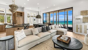 A modern living room with a large window overlooking the ocean, a kitchen with wooden cabinets, and a dining area with a view. - Gulf-Front Luxury Home w/ Panoramic Views • Private Pool • Carillon Beach (Panama City Beach)