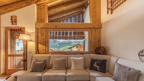 A cozy living room with a wooden ceiling, a sofa with cushions, a window with a view of a mountain, and a lamp. - Casa Verocip, a panoramic attic with a view of the Dolomites (Cortina d´Ampezzo)