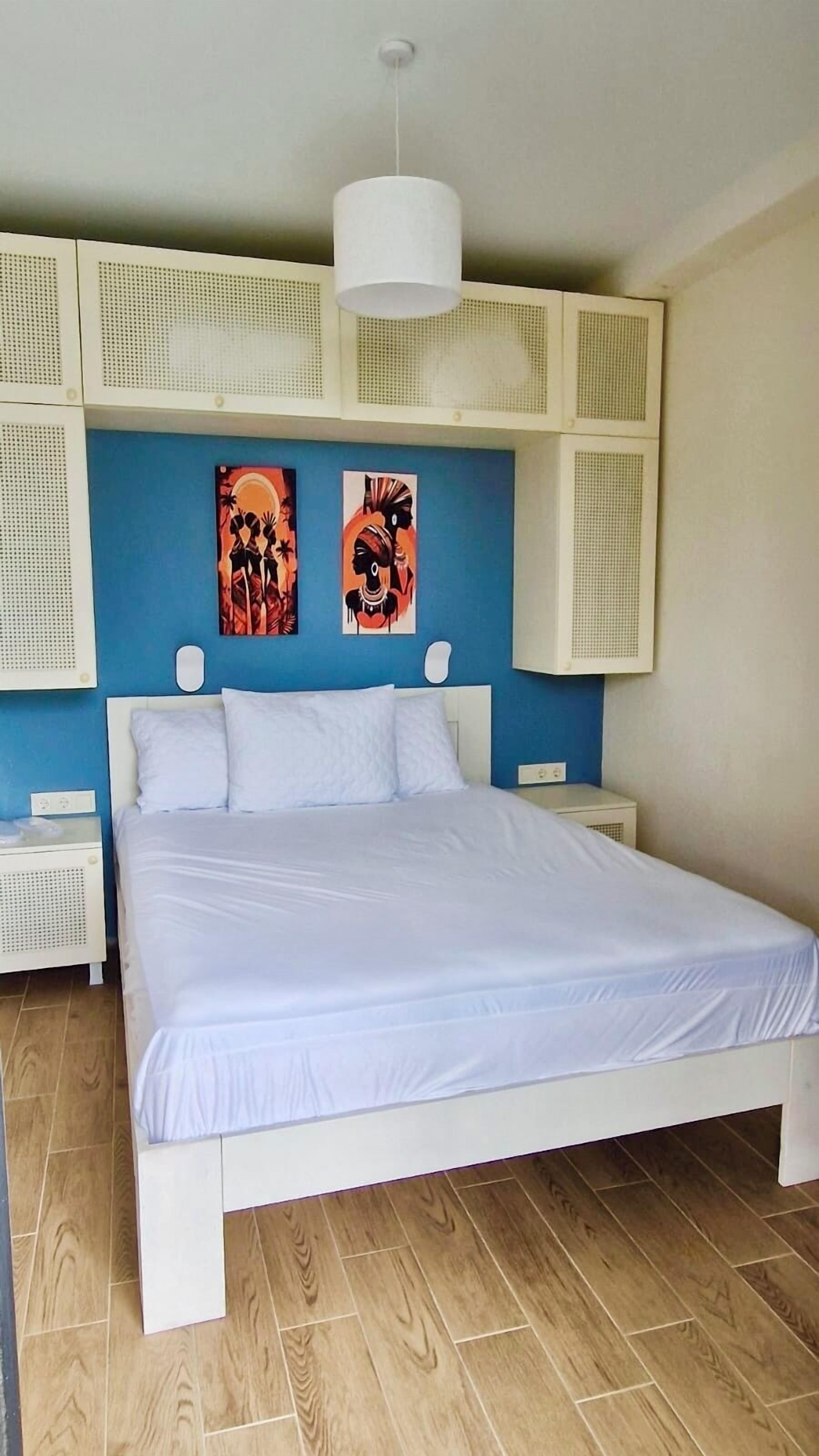 A bedroom with a large bed, two wall-mounted artworks, a white ceiling light, and a white bedside table.