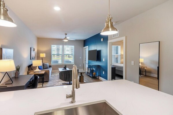 A modern kitchen with a central island, a dining area with a table and chairs, a living room with a sofa and a TV, and a large window with a view of the outside.