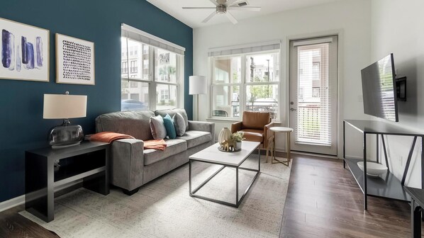 A modern living room with a grey sofa, a coffee table, and a TV on a stand. There are large windows with blinds, a ceiling fan, and artwork on the walls.