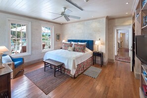 Un dormitorio con cama, ventilador de techo, banco, mesita de noche, televisor y vistas al exterior.
