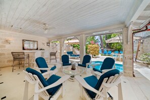 A covered outdoor seating area with white chairs and a glass table, a stone wall, and a pool in the background.