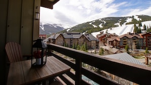 A wooden deck with a table and a lantern, overlooking a ski resort with buildings and snow-covered mountains. - Pp506 Passage Point 1 Bedroom Condo (Copper Mountain)