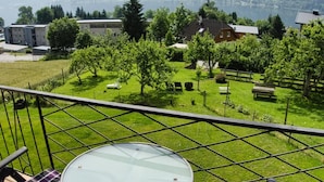A balcony with a glass table and chairs, overlooking a well-maintained garden with trees and a lake view. - Apartment With Mountain View (Treffen)