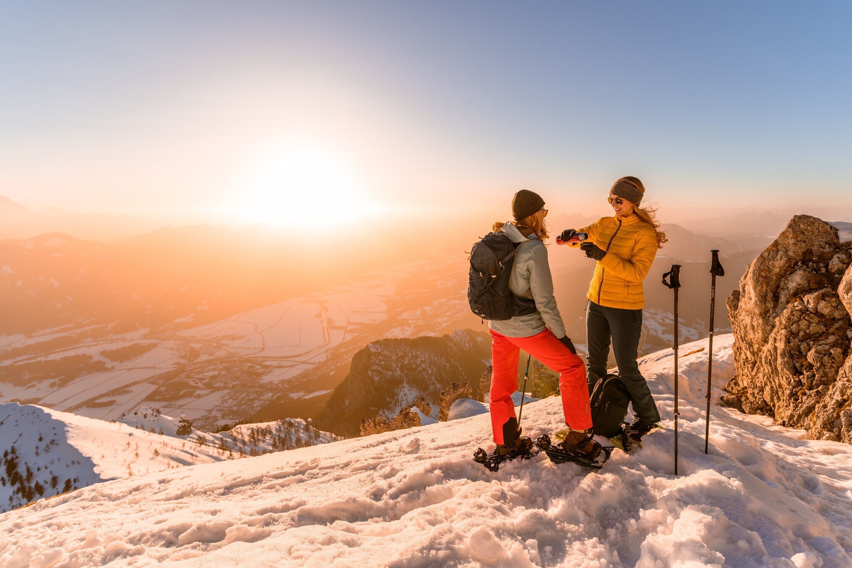 Two people standing on a snowy mountain peak with a clear sky and distant landscape.