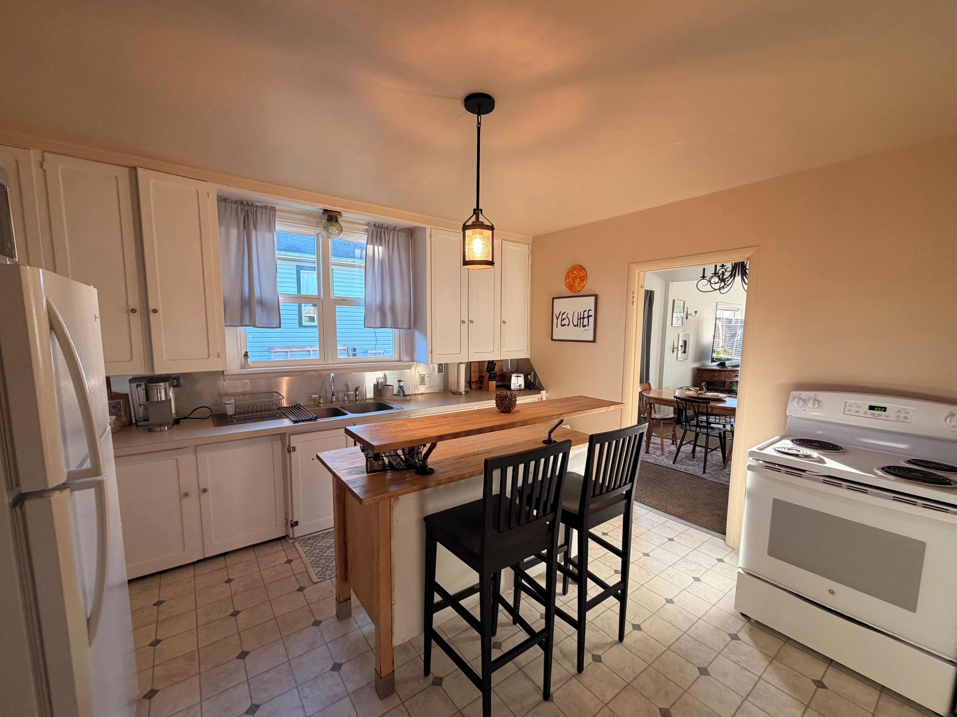 Une cuisine avec des meubles blancs, un îlot central en bois, des chaises noires, une cuisinière blanche et un luminaire suspendu.