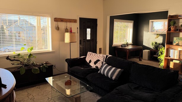 Ein Wohnzimmer mit einem schwarzen Sofa, einem Glastisch und einem Bücherregal. Zwei Fenster mit Jalousien, eine Tür und ein kleiner Tisch mit einer Pflanze sind ebenfalls zu sehen.