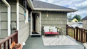 Eine Holzterrasse mit Bank und kleinem Tisch, eine Veranda mit Tür und Blick auf die Straße.