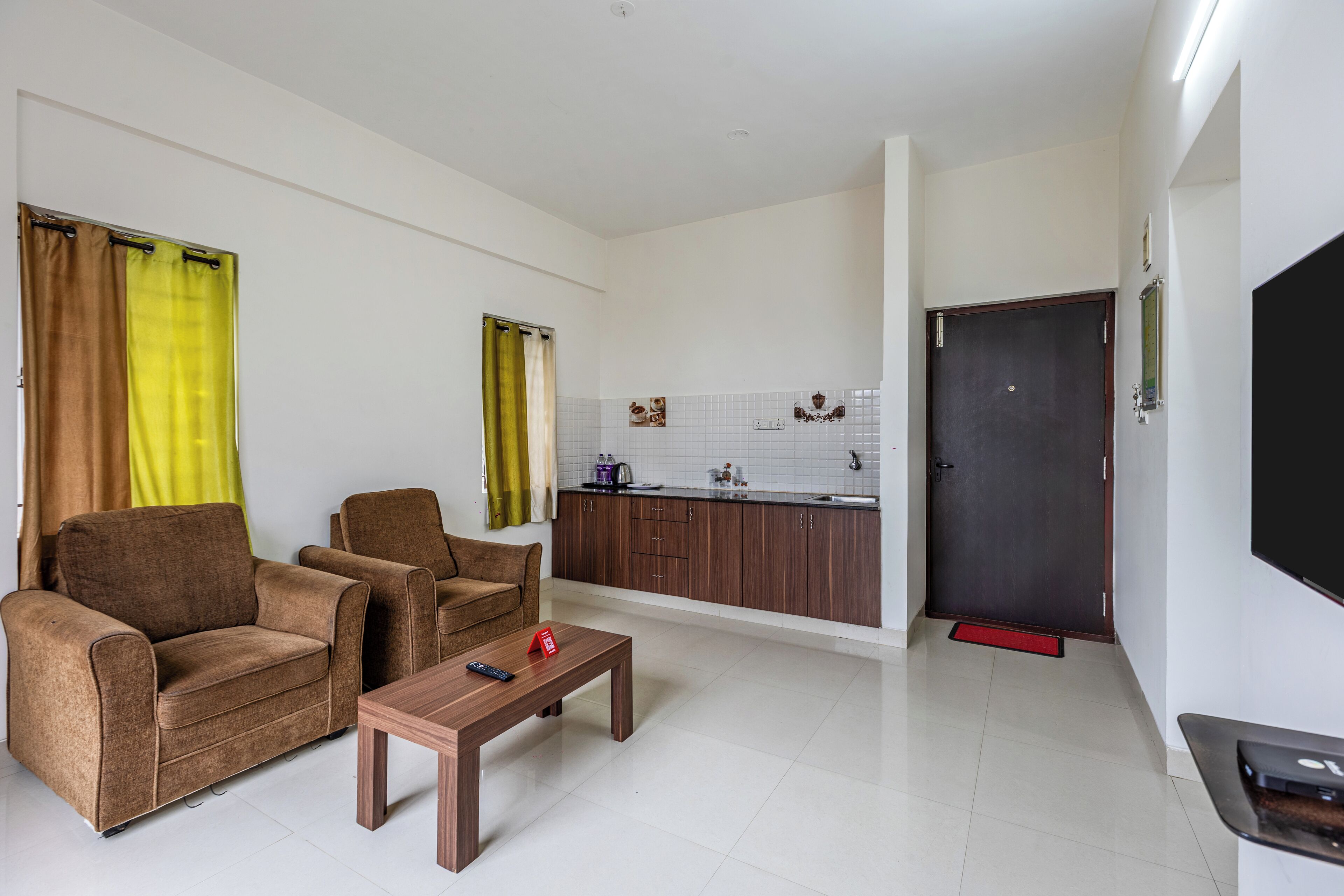 A living room with two sofas, a coffee table, and a TV. There is a kitchen area with cabinets and a door.