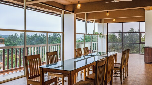 A spacious dining area with a glass table and wooden chairs, large windows offering a view of greenery, and a ceiling fan. - Hotel Acacia Yelahanka (Bengaluru)