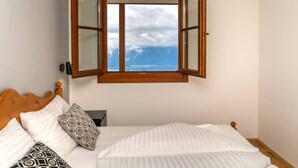 Uma cama com cabeceira de madeira, roupa de cama branca e almofadas estampadas. Uma janela com molduras de madeira oferece vista do céu e das nuvens.