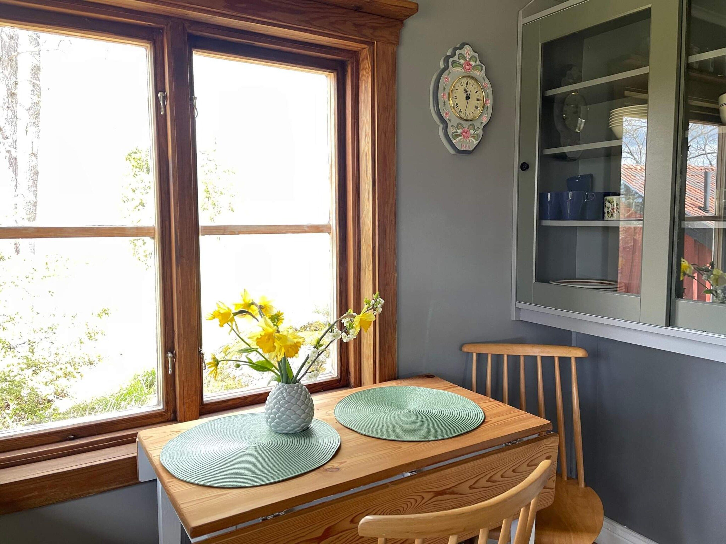 A dining area with a wooden table, two green placemats, a pineapple vase with yellow flowers, and a wall clock.