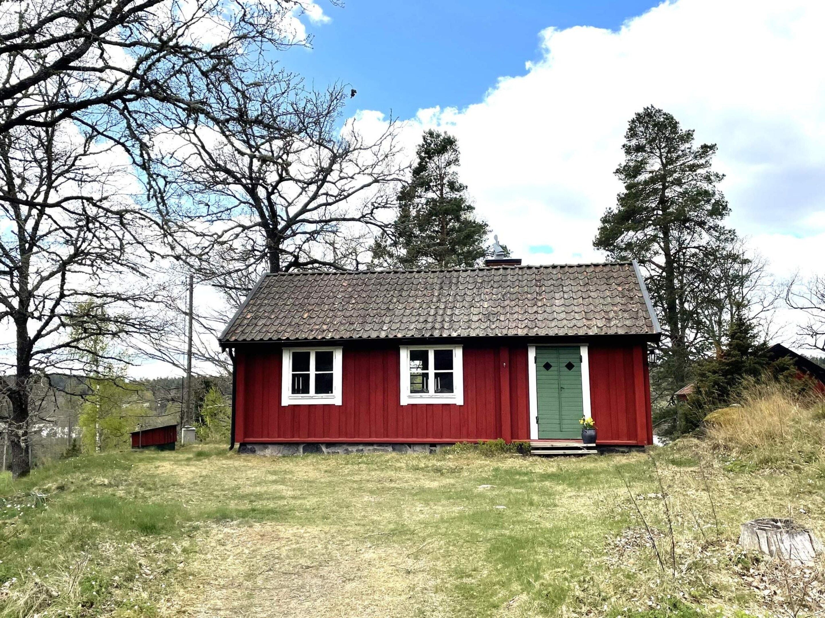 A small red wooden house with a green door, surrounded by trees and grass.