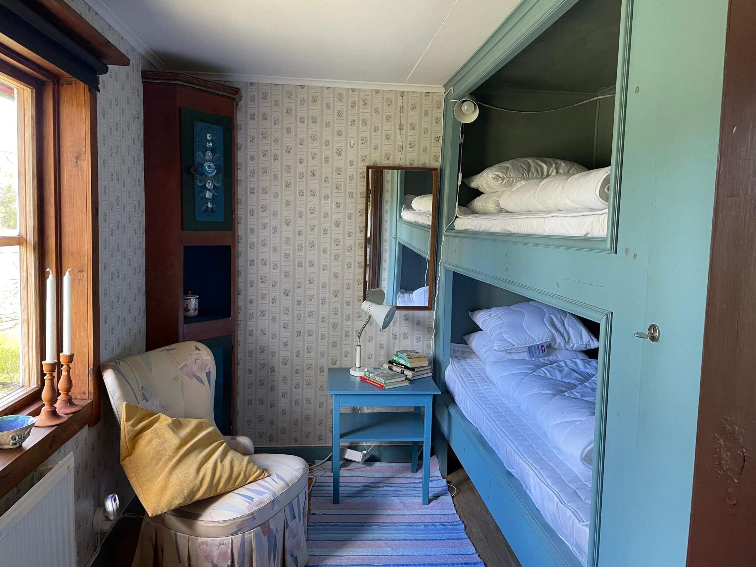 A small room with bunk beds, a chair, a small table with books, a radiator, and a window with curtains.