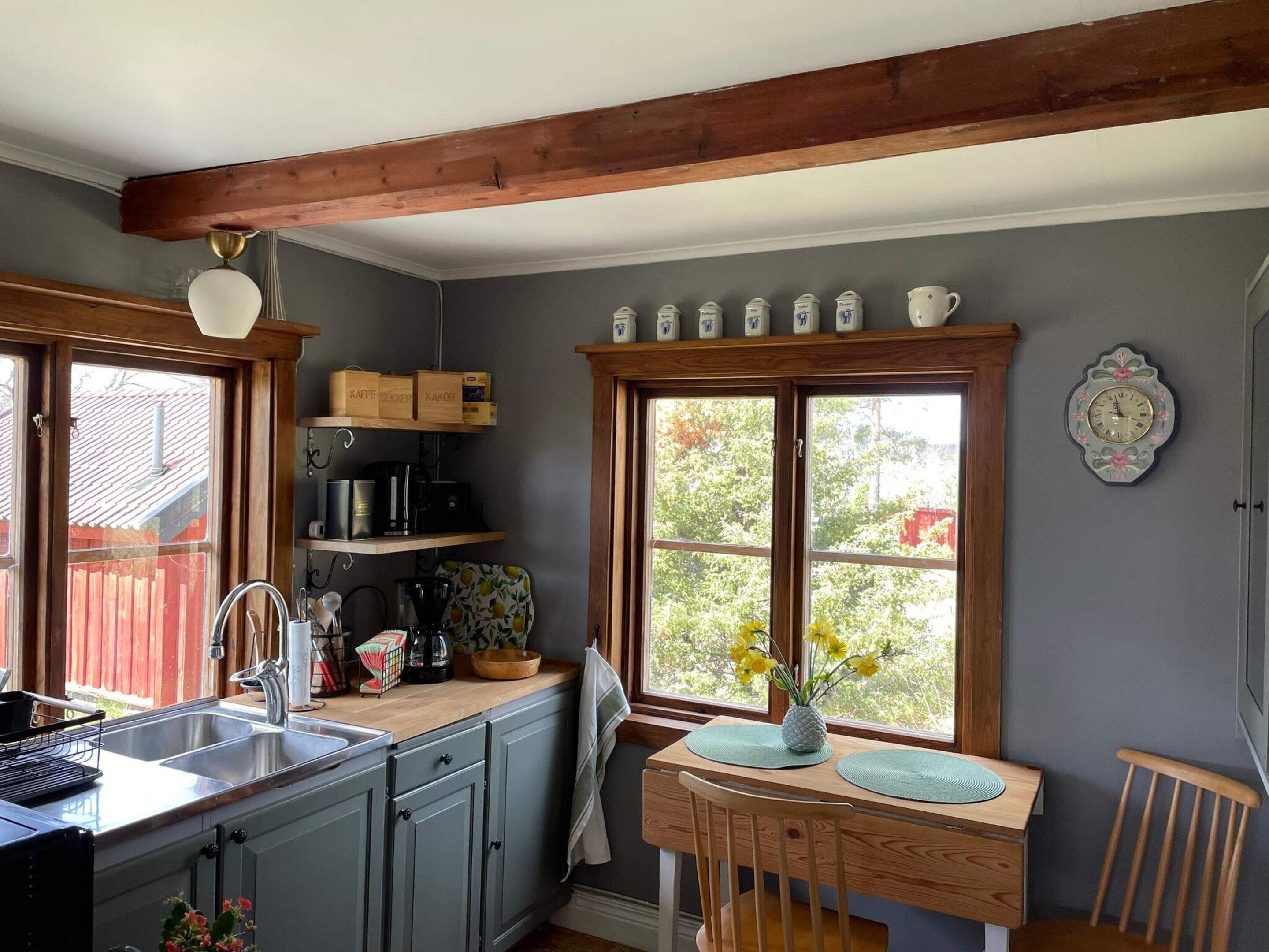 A kitchen with a wooden beam ceiling, a sink, cabinets, a window with a view, and a clock on the wall.