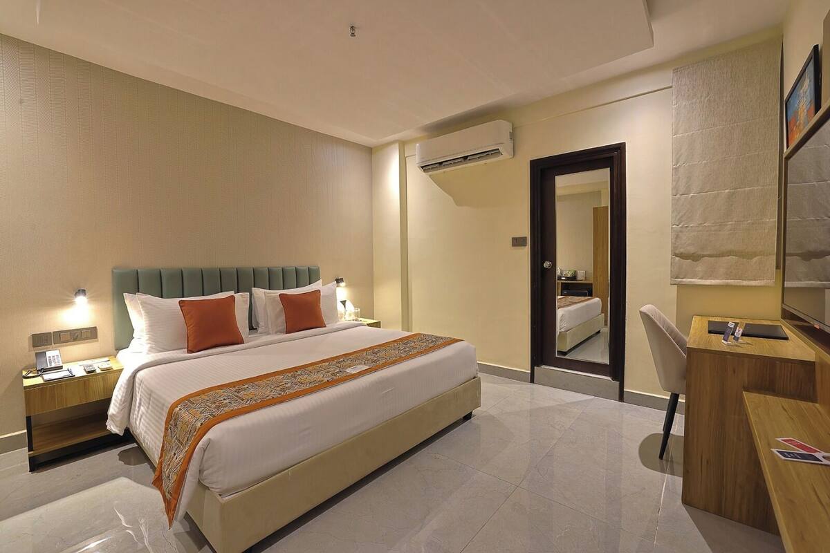 Una habitación de hotel con cama, mesitas de noche, escritorio, silla, aire acondicionado y espejo.