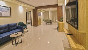 A hotel lobby with a blue sofa, a small table, a TV, and a dining area with chairs and a table.