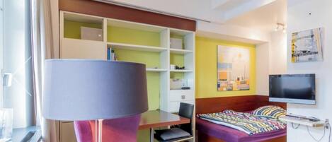 A modern hotel room with a bed, a desk, a chair, a TV, and a window.