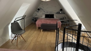 A modern loft bedroom with a sloped ceiling, a bed with a pink blanket, a chair, a small table, and a unique chandelier.