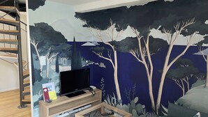 A living room with a TV on a wooden stand, a glass coffee table, and a mural of trees on the wall.