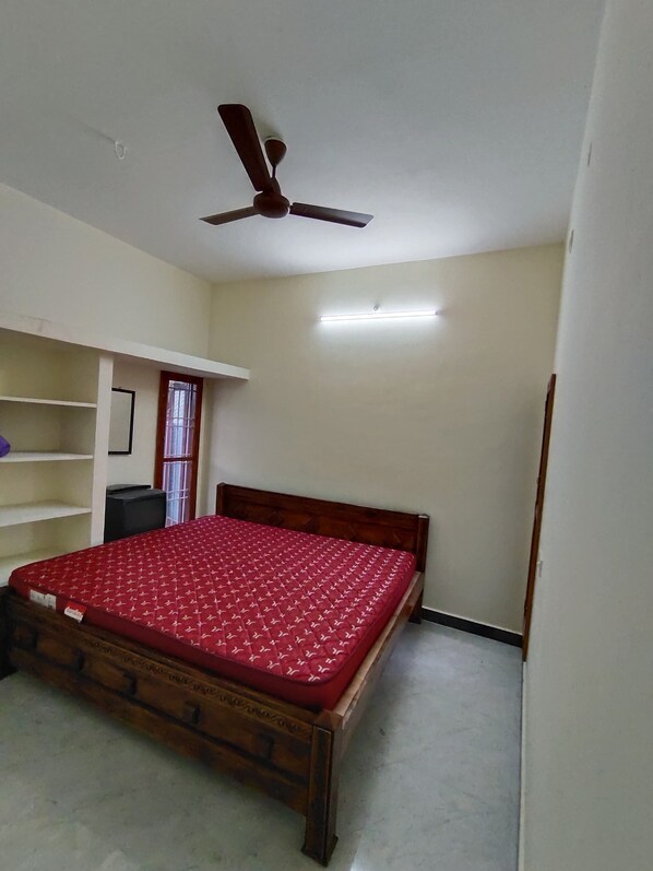 Un dormitorio con cama de madera, ventilador de techo, estantería y ventana con cortina.